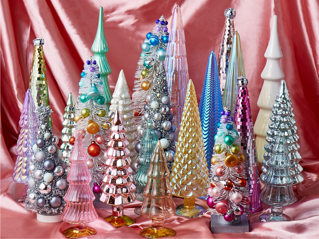 A collection of colorful, decorative Christmas trees made of glass, tinsel, and ornaments is displayed against a pink satin backdrop. The trees feature sparkly, iridescent, and metallic finishes.