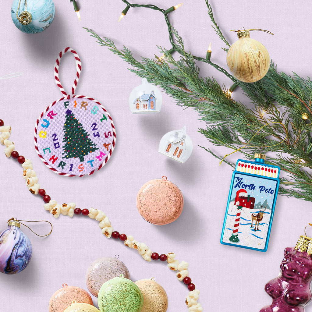 An assortment of ornaments, including painted baubles, macarons, and a stitched “Our First Christmas” design, all against a lavender background with lights, a popcorn garland, and a branch of greenery.